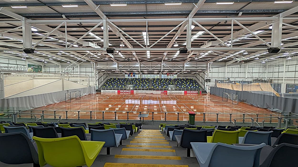 A large indoor sports arena with a polished wooden floor marked for various sports. In the foreground, there are rows of multicoloured plastic seats in blue, green, and black. The background features a tiered seating area with more multicoloured seats. The ceiling is high with exposed beams and bright lighting. Nets hang along the sides of the court, and banners are visible on the far wall.