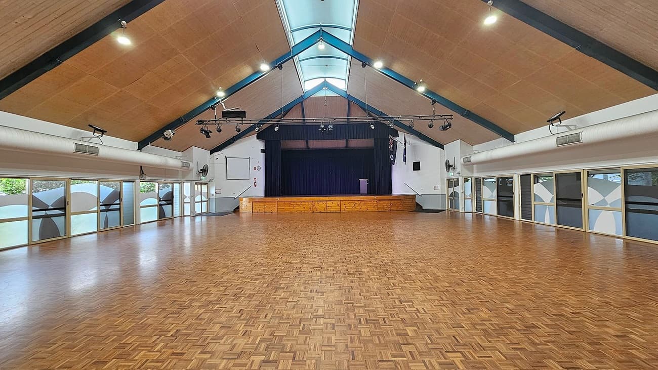 A spacious hall with a high, triangular wooden ceiling and parquet flooring. At the far end, there's a stage with a wooden front and dark curtains. The walls are lined with large windows featuring frosted glass with geometric patterns. Ceiling lights and air ducts are visible, and there are speakers and a projector mounted above. The room is empty, providing an open, unobstructed view of the entire space.