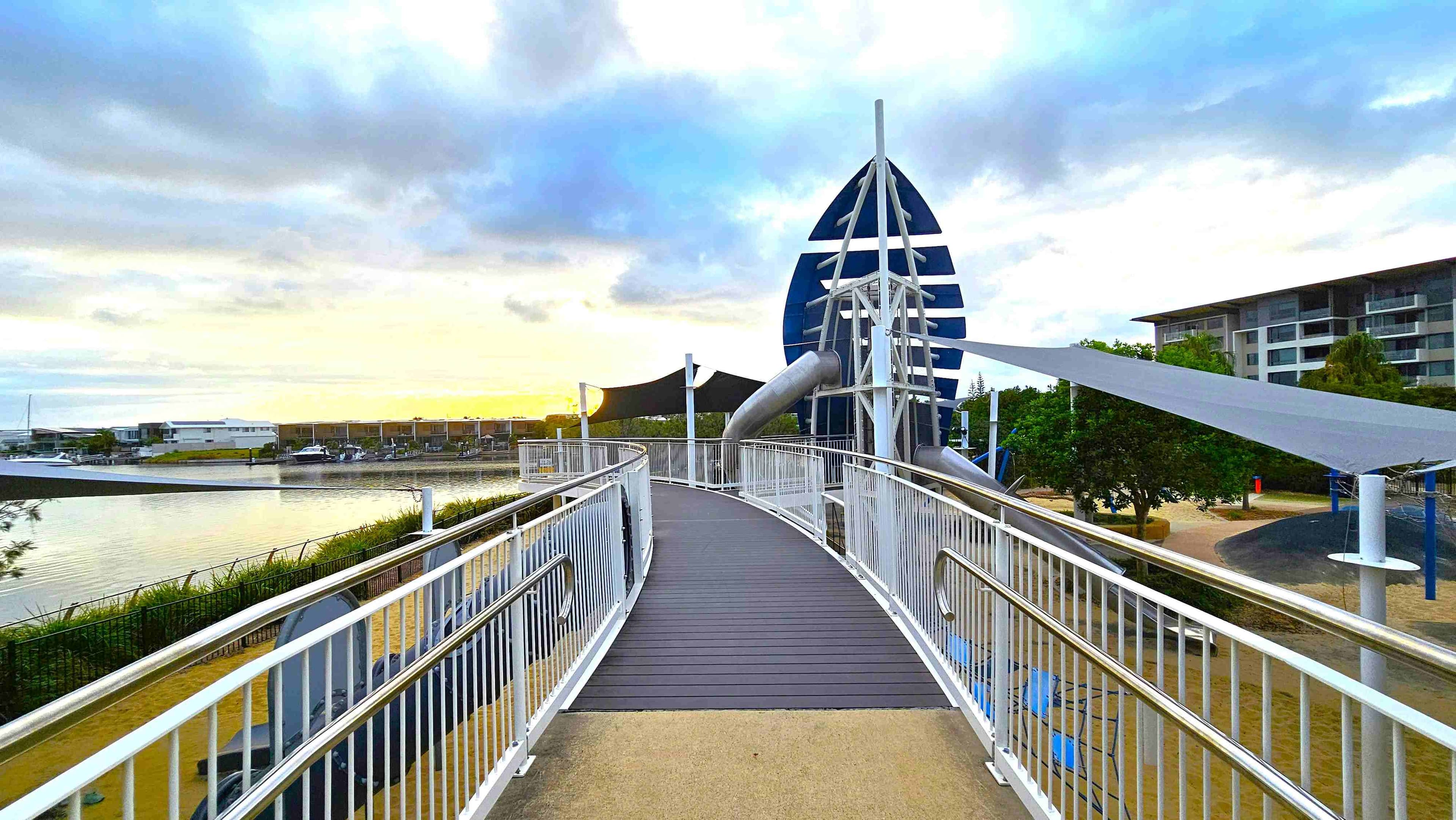A playground with a sandy surface and a path winding through it. On the left, there are shade sails over a climbing structure with ropes and blue poles. In the centre, a mound with blue dome-shaped climbing features. On the right, a tall blue spiral slide and swings. Trees are scattered throughout, and a modern white building is visible in the background. The ground is a mix of sand, rubber, and concrete.