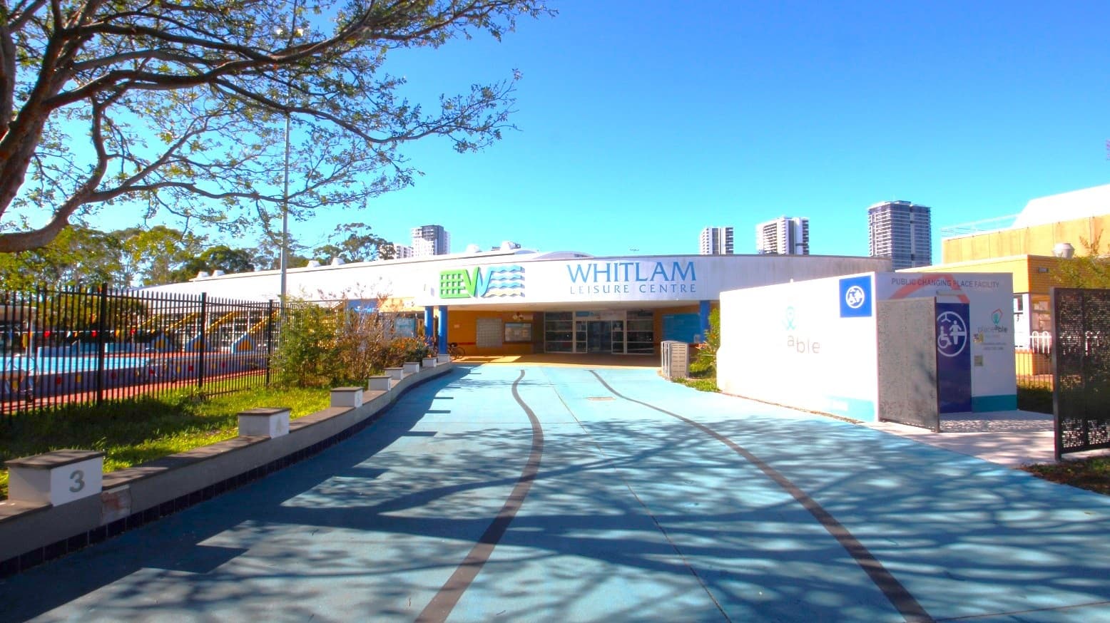 A smooth grey concrete pathway leading to the Whitlam Leisure Centre. The venue is a low-set building with grey, white, and blue painting. Upper wall of the front face of the building shows the name of the venue and its logo. On the left is a 50-meter external pool secured by a black fence, while on the right is a public changing place facility by Placeable Solutions. The entry point to the facility is through a black gate.