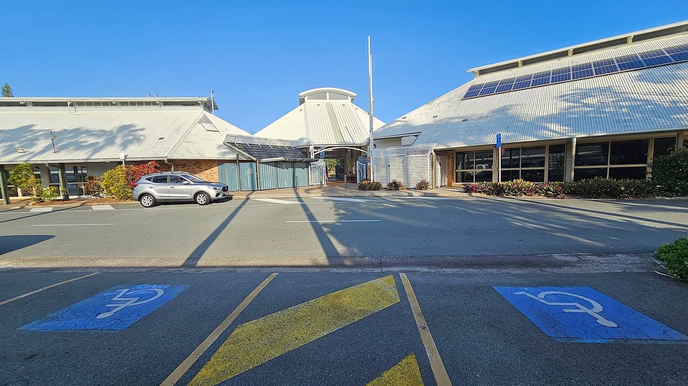 A car park with two marked disabled parking spaces in the foreground. A silver car is parked on the left side near a building with a sloped roof. The building has solar panels on the roof and is surrounded by plants. In the centre, there is a covered walkway leading to an entrance. The right side features another building with large windows and more solar panels. Shadows of trees are cast on the ground and buildings.