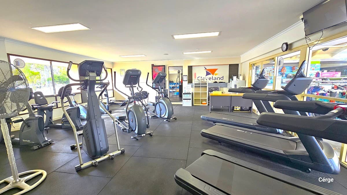 Free weights and resistance exercise machinery at Cleveland Aquatic Center. Similar to the cardiovascular section of the gym, the room has a grey tile floor. It also has glass windows on the back white wall. The room also has a white ceiling but with longer, narrower, rectangular fluorescent lights.
