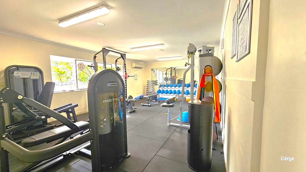 Free weights and resistance exercise machinery at Cleveland Aquatic Center. Similar to the cardiovascular section of the gym, the room has a grey tile floor. It also has glass windows on the back white wall. The room also has a white ceiling but with longer, narrower, rectangular fluorescent lights.
