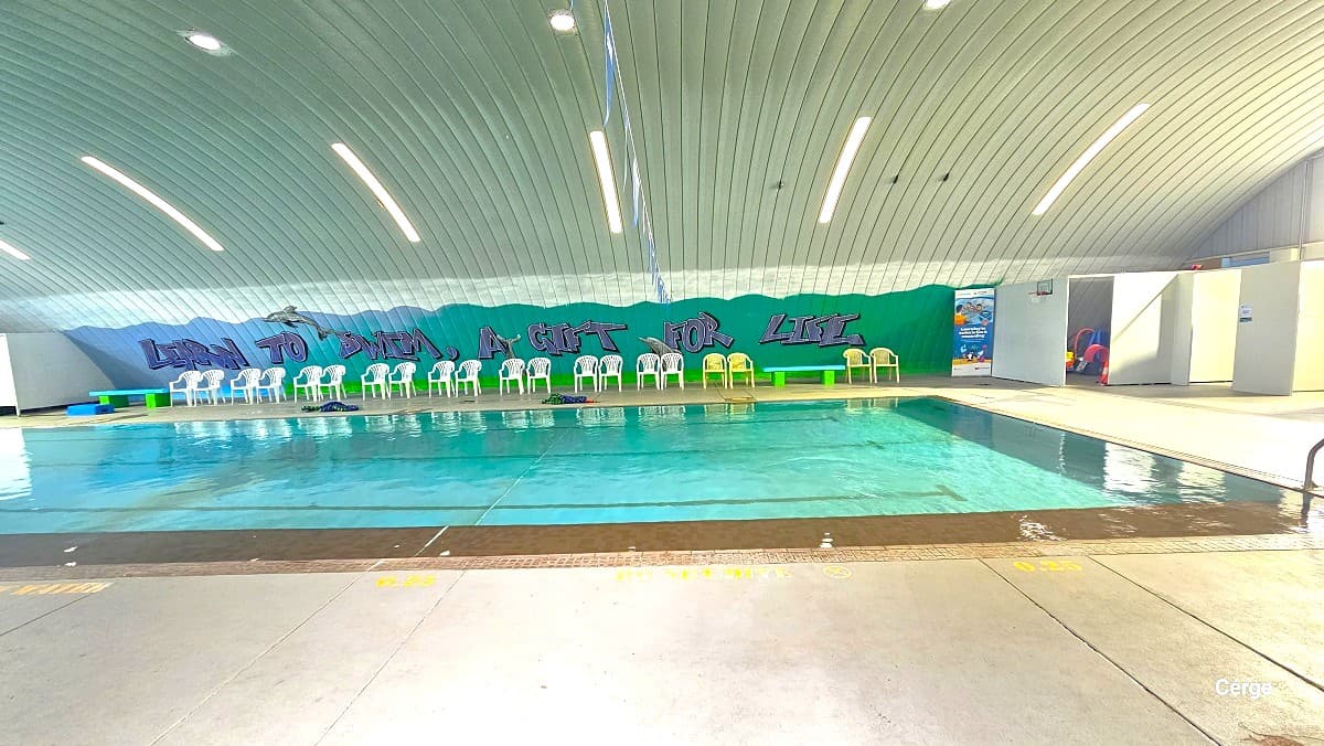 View of the indoor pool at Cleveland Aquatic Centre. The depth of the water is indicated next to the brown tiled pool wall which is 0.25 meter. A NO DIVE phrase is detailed in yellow also to the pool side. There is a blue bench and white plastic chairs on the opposite side of the pool, next to the green wall with graffiti that says, “Learn to Swim, A Gift for Life”. The ceiling has a corrugated iron roof dome pattern.