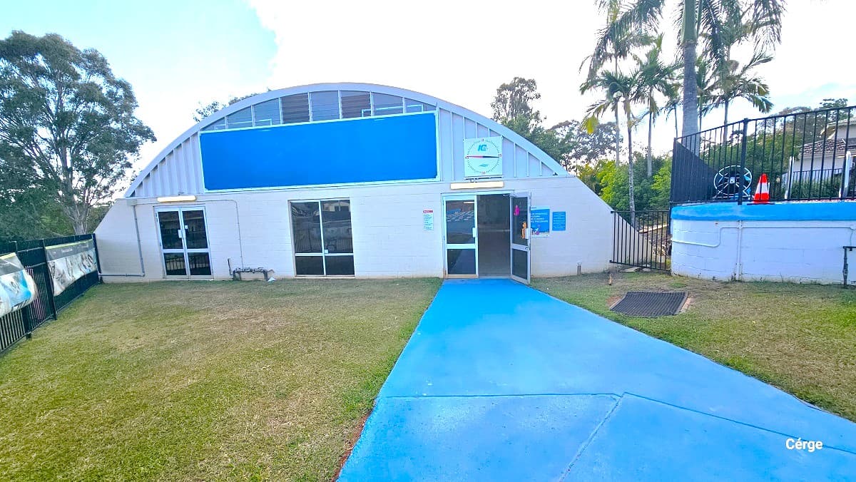 Entrance to the indoor pool at Cleveland Aquatic Centre. The indoor pool has a dome-like building structure and a combination of cream colored brick wall and a corrugated upper iron wall. It has three manually operated glass doors lined with white metal frames. The cemented pathway leading to the right door is painted in blue. At the upper part of the dome is a large blue rectangular shape. The perimeter is enclosed by as black vertical bar fence.