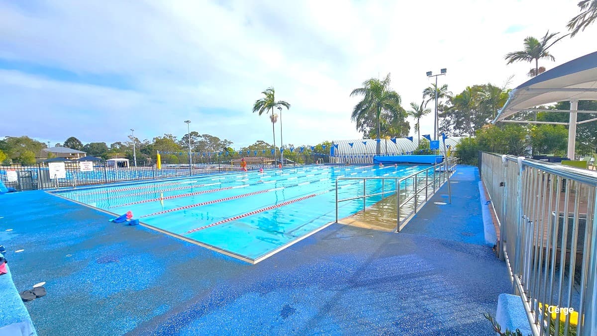 The 25m six lane pool at the outdoor aquatic area has ramp access with handrails on the right pool side. The pool deck has blue color floor markings. This pool is separated from the other pool and from the reception area by handrails enclosing the entire pool perimeter. Each lane is separated by red, white, blue, and floating rope lanes. Strings of triangular blue flags are wrapped around the opposite poles of the pool with a yellow floating device.