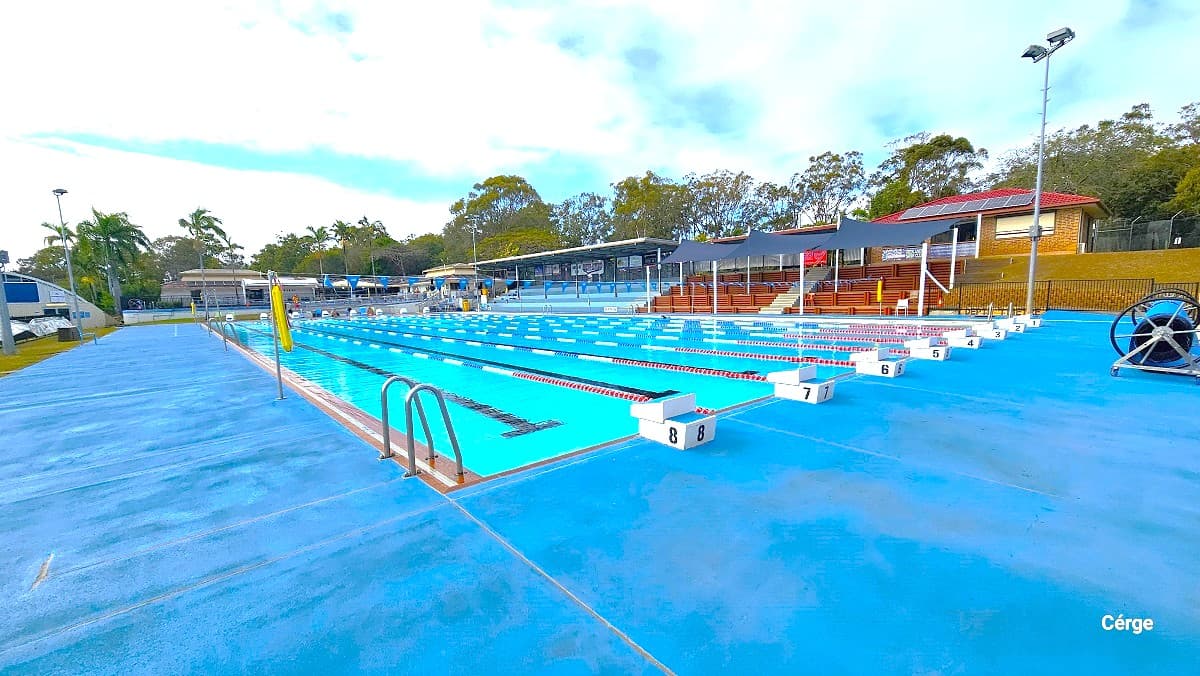 Cleveland Aquatic Centre has a 50m outdoor pool with a wide pool deck. On the right side is brown-tiered seating with a black sunshade and blue-tiered seating with a solid roof. Behind it are low-set brick cream-coloured buildings with brown tiled roofing having solar panels. Pool lanes are marked by white diving blocks marked from 1 to 8 and are separated by red, blue, and white floating rope lanes.