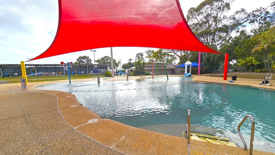 Closer look at the Splash pool. There is a yellow pool ladder with black edgers and metal handrails. A fast-flowing current coming from a metal bollard in the center splashes into the pool water. There is a blue tent next to a white mushroom-like hole after the tall spraying fountains. A number of chairs are placed against the corrugated wall at the back.