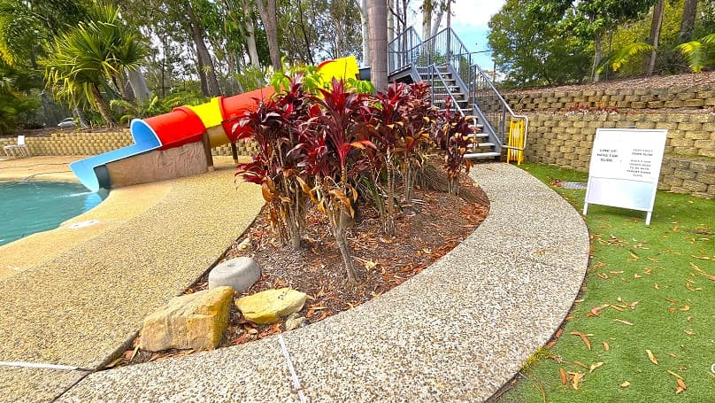 A natural stone pathway connects to the stairs for the yellow and orange spiraling waterslide. A small garden that separates the pool deck and the pathway has bright red leaf plants. There is a white signboard on the right side of this pathway. A stone wall separates the pool deck and upper garden area with trees, shrubs, and bushes.