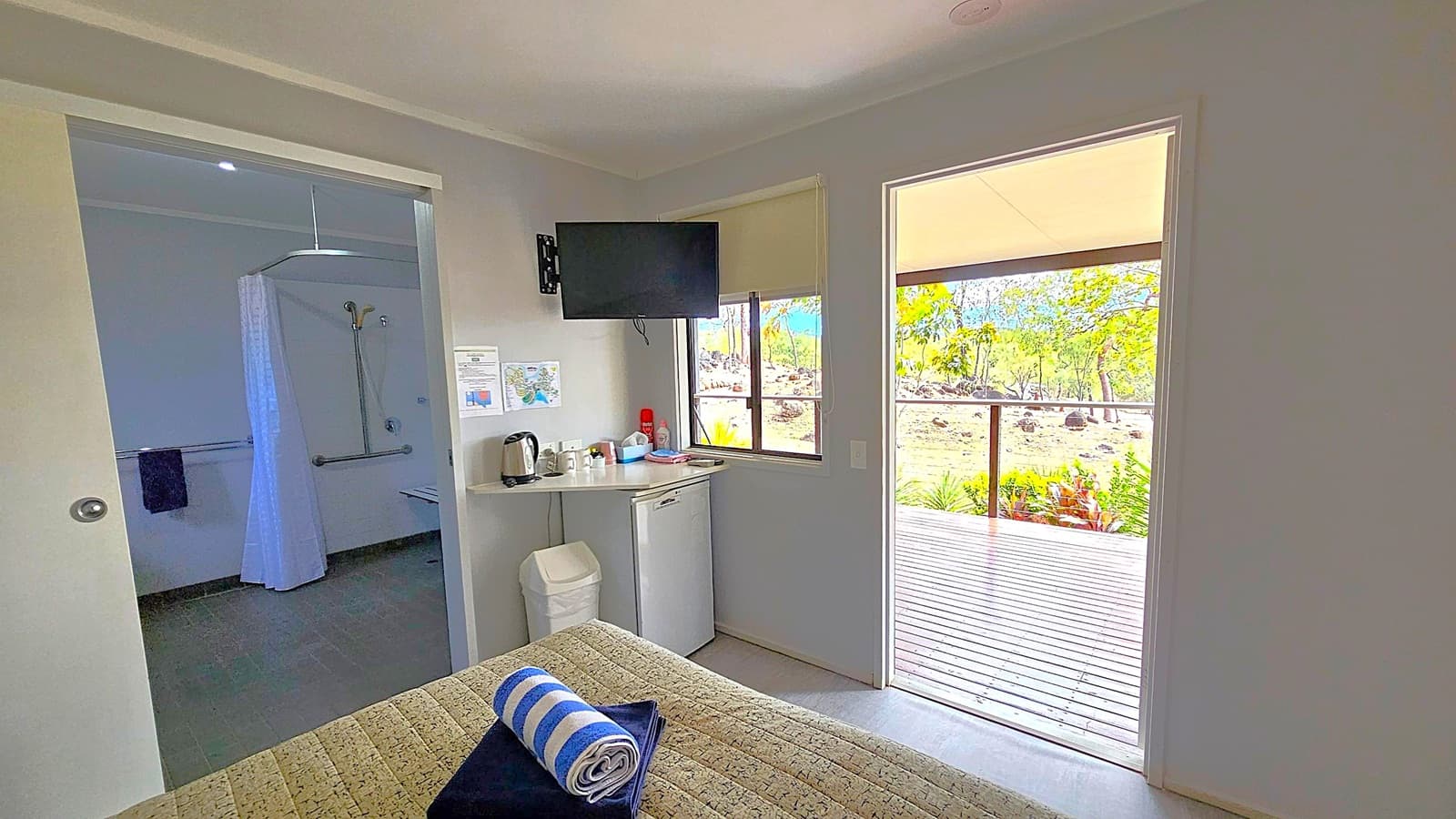 A room with a view of a wooden deck and greenery outside. On the left, there's an open door leading to a bathroom with a shower and curtain. In the centre, a small counter holds a kettle and cups, with a mini fridge below. A wall-mounted TV is above the counter. A bed with a patterned cover and a rolled towel is in the foreground. The walls are light-coloured, and the ceiling is plain.