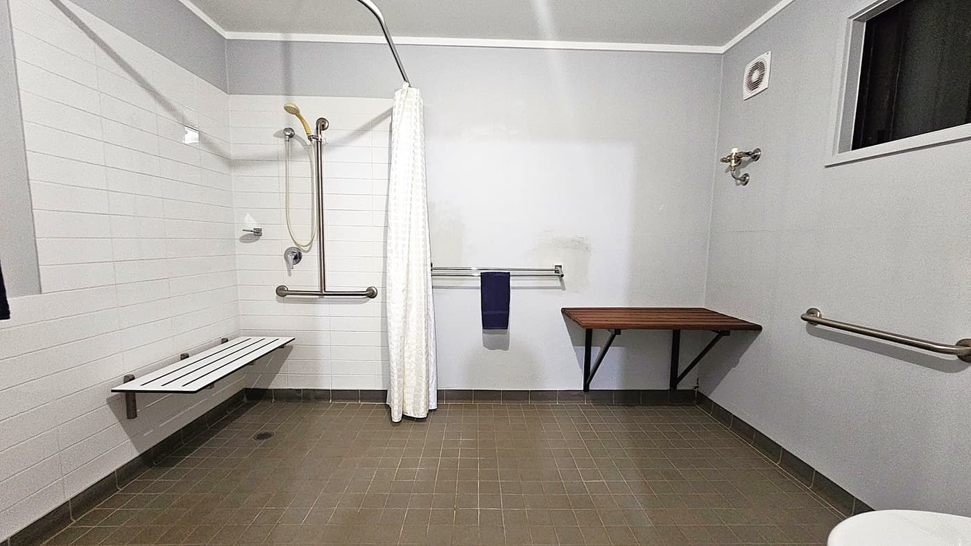 A bathroom with a green tiled floor and white walls. On the left, there is a shower area with a white curtain, a handheld showerhead, and a foldable bench. A grab bar is mounted on the wall. In the centre, a towel rail holds a dark towel. On the right, a wooden shelf is attached to the wall, and a small window is above it. A grab bar is also visible on the right wall. The ceiling is plain white.