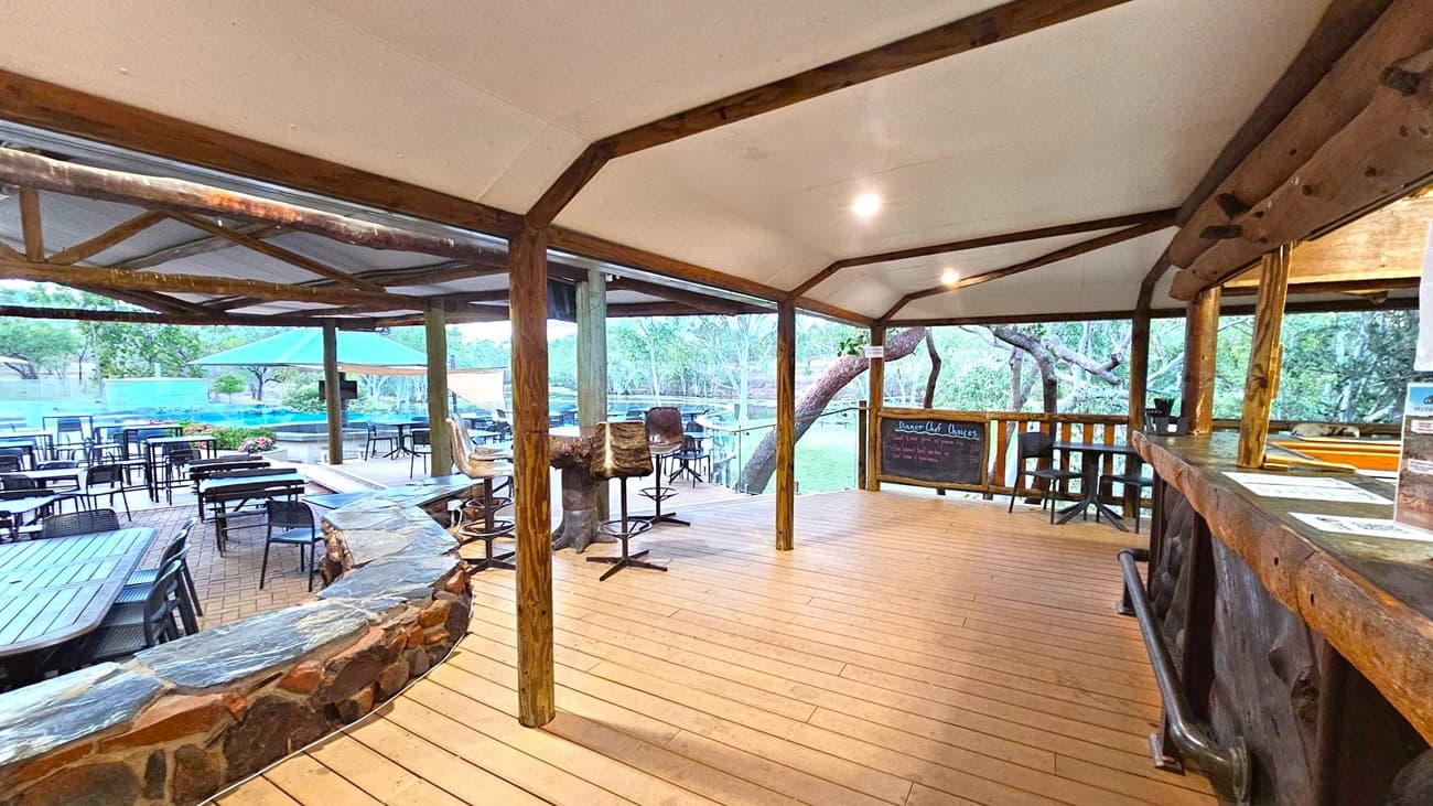 A rustic outdoor dining area with wooden flooring and a wooden ceiling supported by timber beams. On the right, there's a bar counter with menus and a chalkboard listing dessert options. In the middle, high stools with fur covers are placed around a table. On the left, several tables and chairs are arranged on a brick patio, with a stone wall separating the spaces. In the background, a large umbrella provides shade, and trees are visible beyond the patio.