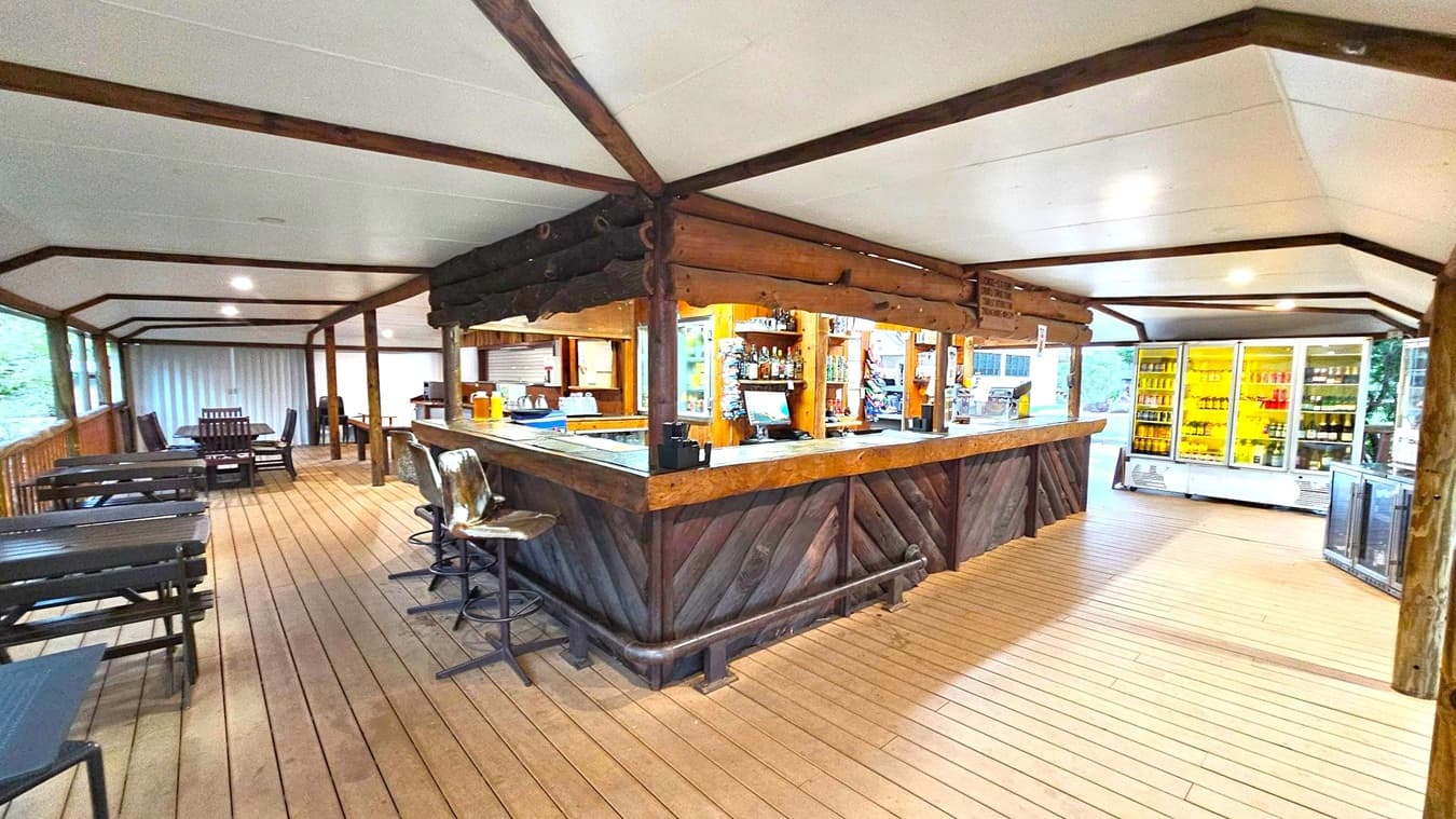 A rustic bar area with a wooden counter in the centre, surrounded by high stools. Behind the bar, shelves are stocked with various bottles and snacks. To the right, there are large refrigerators filled with drinks. On the left, several tables and chairs are arranged along the wooden floor. The ceiling is white with wooden beams, and the space is well-lit with recessed lighting. The background shows a view of greenery through large windows.