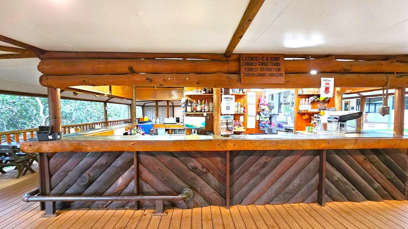 A rustic bar with a wooden counter and diagonal wood paneling. Behind the bar, shelves hold various bottles and snacks. A sign with trading hours is visible above. The ceiling is white, supported by wooden beams. The floor is made of wooden planks. To the left, there are outdoor seating areas with wooden tables and chairs, surrounded by greenery. The right side shows more shelves and a cash register.