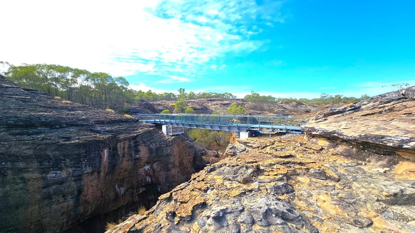 A rugged landscape with a metal bridge spanning a deep gorge. The foreground features rocky terrain with layers of exposed stone. The bridge, with railings, crosses from left to right, connecting the rocky outcrops. In the background, sparse trees and shrubs are scattered across the rocky hills. The sky is mostly clear with some clouds.