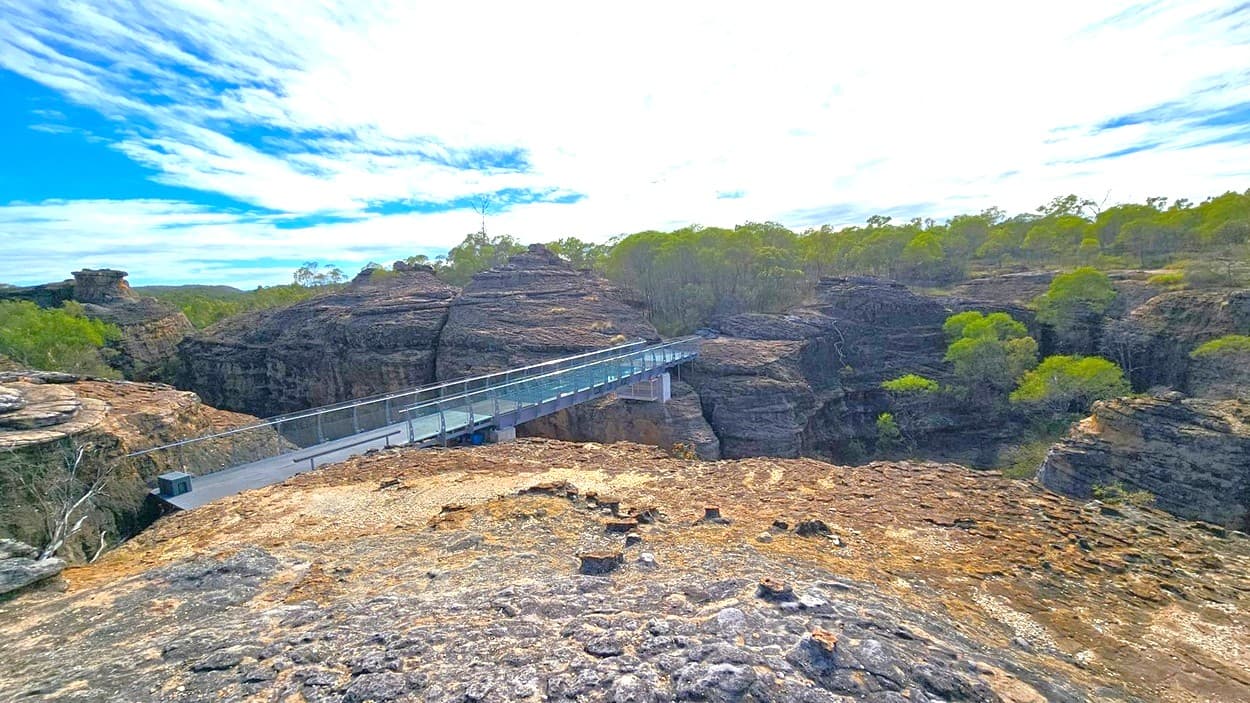 A rocky landscape with a modern glass and metal bridge extending across a deep gorge. The foreground features rugged, uneven rock surfaces with sparse vegetation. In the background, dense green trees cover the tops of the rock formations, creating a natural canopy. The bridge connects two large rock formations, providing a pathway over the chasm.