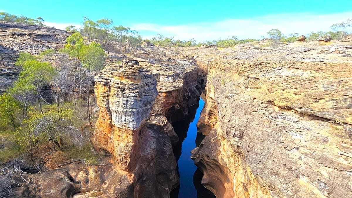 A narrow gorge with steep, rocky walls stretches through the centre, with a small stream of water visible at the bottom. The rock formations are layered and rugged, displaying shades of brown and grey. Sparse vegetation, including small trees and shrubs, grows on the rocky terrain on both sides of the gorge. The landscape is arid, with a clear blue sky above.
