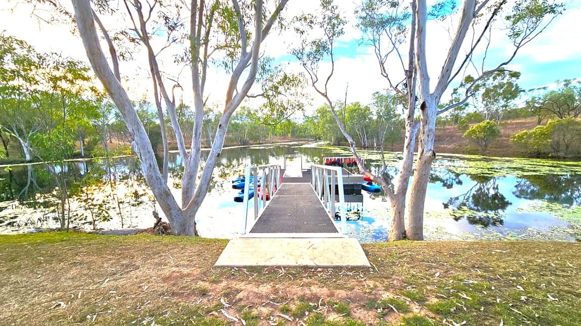 A narrow metal walkway extends from the foreground to a floating dock on a calm lake. The dock is surrounded by several kayaks and paddle boats. Tall trees with slender trunks and sparse foliage frame the scene on both sides. The water reflects the trees and sky, with patches of green lily pads scattered across the surface. The ground in the foreground is a mix of grass and bare earth, with some fallen leaves.