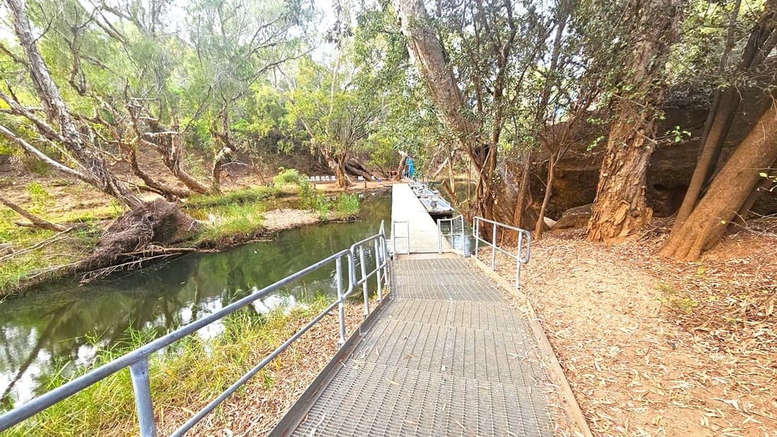 A narrow metal walkway with railings extends over a calm, narrow river. On the left, the riverbank is lined with dense trees and shrubs, their branches extending over the water. On the right, the ground is covered with dry leaves and more trees, with a dirt path running alongside. In the background, the walkway leads to a small dock area with boats moored and a few chairs visible. The scene is surrounded by a natural, wooded environment.