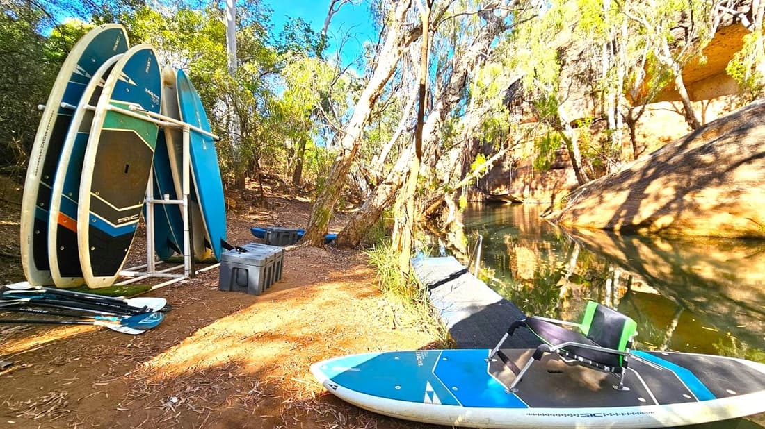 On the left, several paddleboards are stacked vertically on a white rack, with paddles lying on the ground nearby. In the centre, a dirt path is surrounded by trees with a few storage boxes scattered around. On the right, a paddleboard with a seat is positioned on a small dock leading to a calm body of water. The background features a rocky cliff face partially covered by trees, casting shadows on the water.