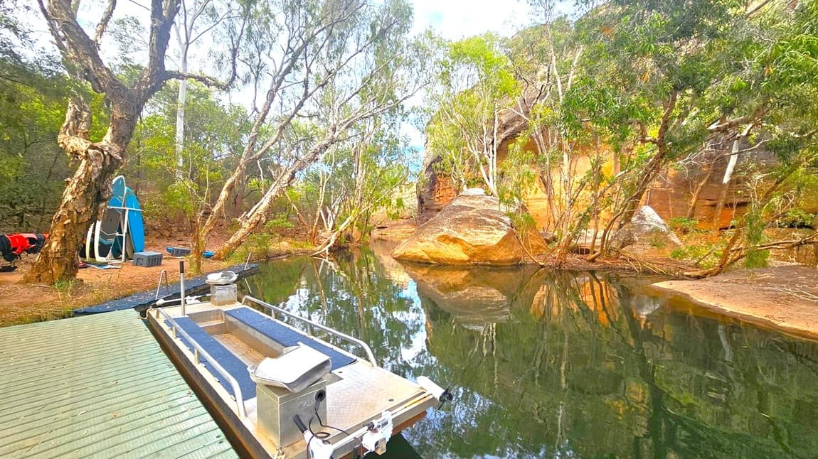 A serene waterway with a small boat docked on a wooden platform in the foreground. To the left, several paddleboards are propped against a tree, with more trees and some equipment nearby. The right side features a large rock formation partially submerged in the water, surrounded by trees. The background is filled with dense greenery and a rocky cliff, with reflections visible on the calm water surface.