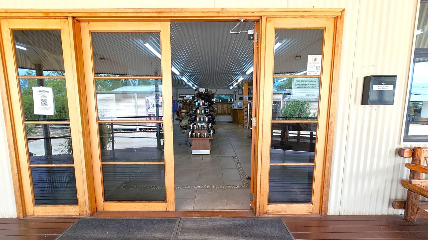Wooden-framed glass doors lead into a shop with a corrugated metal ceiling and tiled floor. Inside, a display stand with various items is centred, surrounded by shelves and counters. Fluorescent lights line the ceiling. Outside, a wooden deck is visible, with a bench on the right. A black box labelled "After Hours Key Return" is mounted on the wall to the right of the door. Signs and notices are posted on the doors.