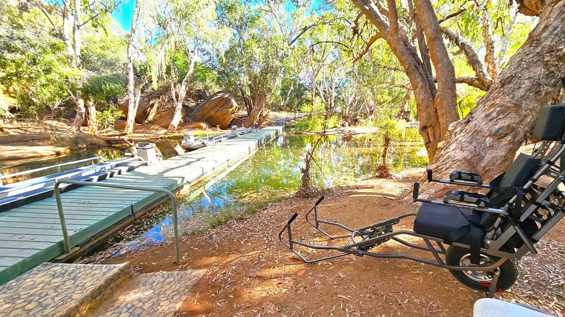 A serene outdoor scene with a wooden dock extending over a calm body of water on the left. The dock has metal railings and is surrounded by lush trees and greenery. On the right, a specialised wheelchair is positioned on a dirt path, next to a large tree with a textured trunk. The ground is covered with dry leaves and the area is shaded by the tree canopy. Steps lead down to the dock, and the water reflects the surrounding foliage.