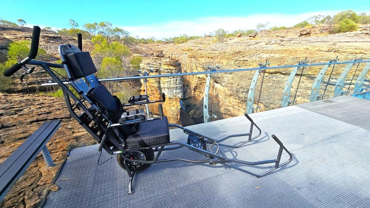 A rugged wheelchair is positioned on a metal mesh platform with a glass railing. The background features a rocky landscape with sparse vegetation and a deep gorge. The platform extends over the edge, providing a clear view of the canyon below. The wheelchair has a sturdy frame and is designed for off-road use. The surrounding area is arid, with scattered trees and shrubs on the rocky terrain.
