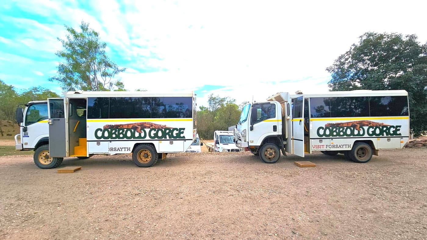 Two white tour buses with "Cobbold Gorge" written on the sides are parked on a dirt surface. The bus on the left has its door open, revealing yellow steps. Both buses have large windows and are facing each other. In the background, there are trees and several other vehicles partially visible. The sky is partly cloudy.