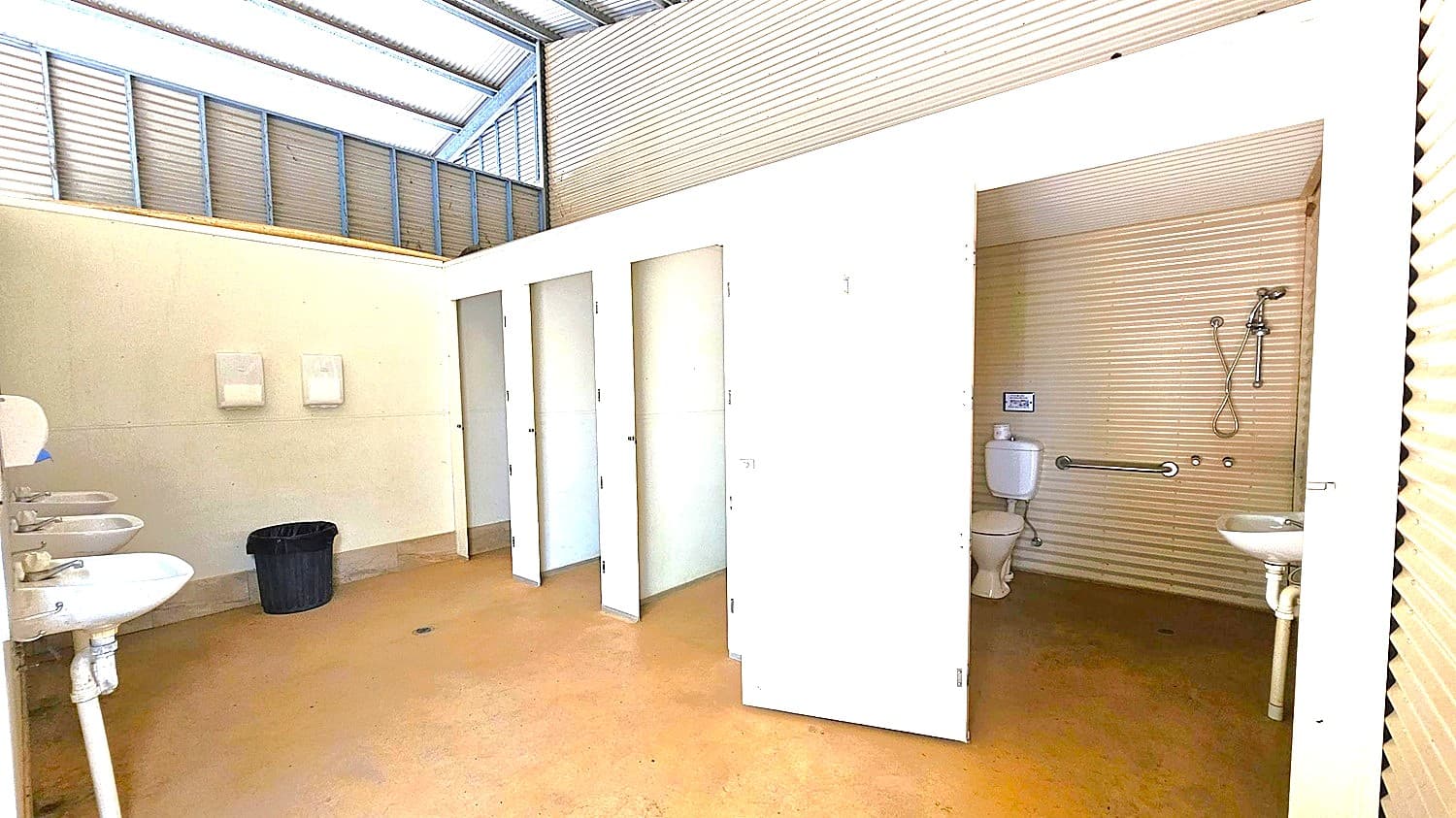 A public restroom with a corrugated metal ceiling and walls. On the left, there are three white sinks with mirrors above them and a black waste bin on the floor. In the centre, there are three closed white stall doors and one open door revealing a toilet, a grab bar, and a small sink. The floor is a light brown colour, and the upper walls have a light green hue. The ceiling has a slanted design with translucent panels allowing natural light.
