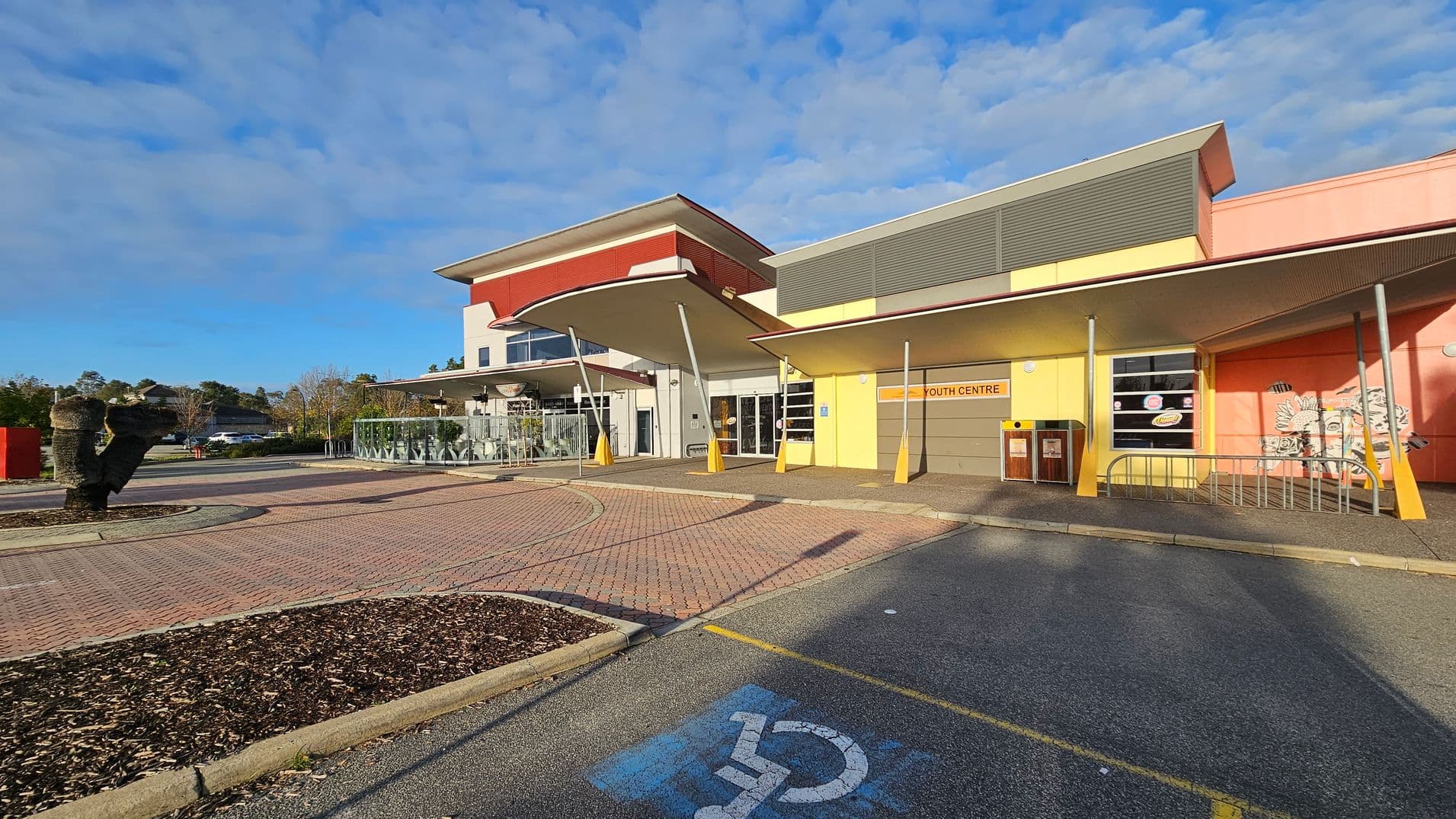 A modern building with a colourful facade, featuring yellow and orange walls, and a sign reading "Youth Centre" above the entrance. The structure has a distinctive overhanging roof supported by angled poles. In the foreground, there is a paved area with a designated disabled parking space marked in blue. To the left, a landscaped area with mulch and a sculptural element. The background includes trees and a clear sky.