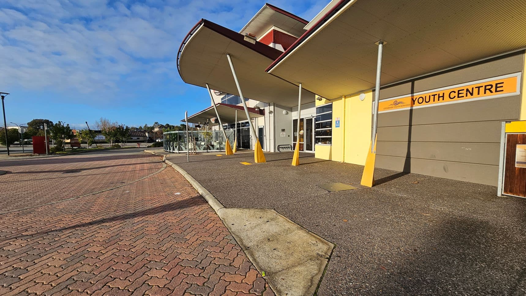 A building with a sign reading "Youth Centre" on the right, featuring a yellow and grey facade. The structure has a modern design with slanted pillars supporting overhanging roofs. The foreground shows a paved area with a mix of concrete and brick patterns. On the left, there are trees and a few street lamps along a pathway. The background includes more trees and distant buildings under a partly cloudy sky.