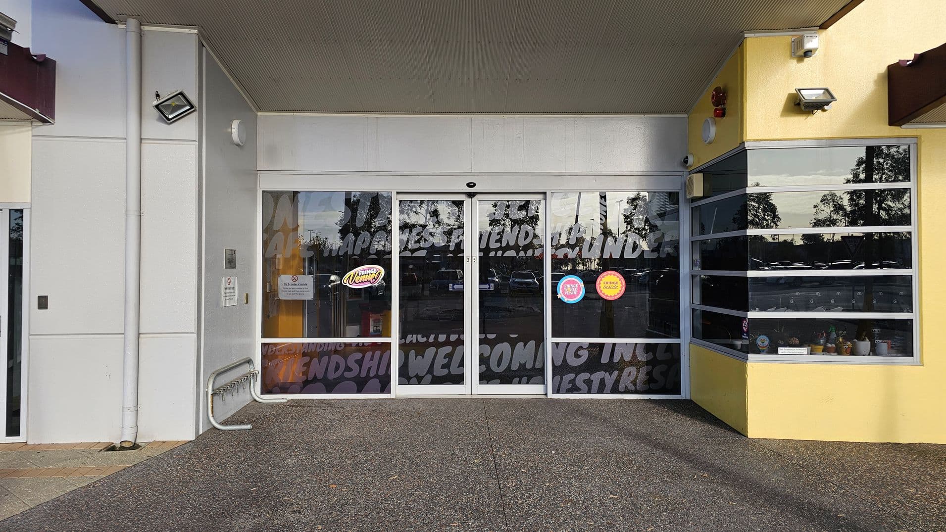 A storefront with large glass sliding doors in the centre, featuring text and logos. To the left, a white wall with a mounted light and a bike rack. On the right, a yellow wall with a window display showing various small items. The ceiling is green, and the floor is a speckled concrete surface.