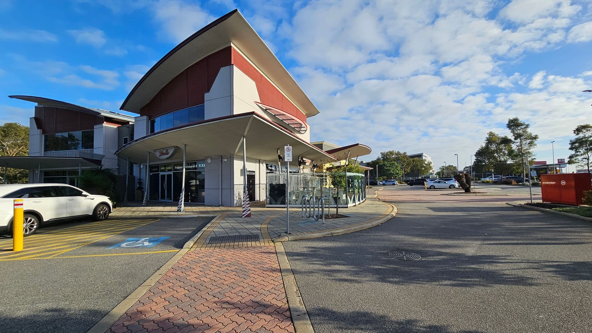 A modern building with a curved roof is on the left, featuring large glass windows and a red accent wall. A white car is parked in a designated disabled parking space with blue and yellow markings. In the centre, there is a bicycle rack and a signpost. The right side shows a spacious car park with a few trees and vehicles. The ground is paved with a mix of asphalt and brick patterns.