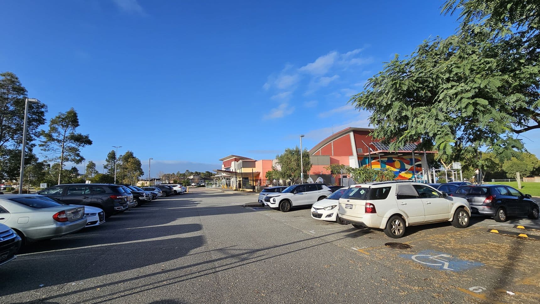 A car park with several vehicles is on the left, including a white SUV in the foreground. On the right, a building with a colourful mural and a red roof is partially visible, surrounded by trees. The ground is paved, and there is a marked disabled parking space in the foreground. Tall streetlights and more trees are in the background under a clear blue sky.