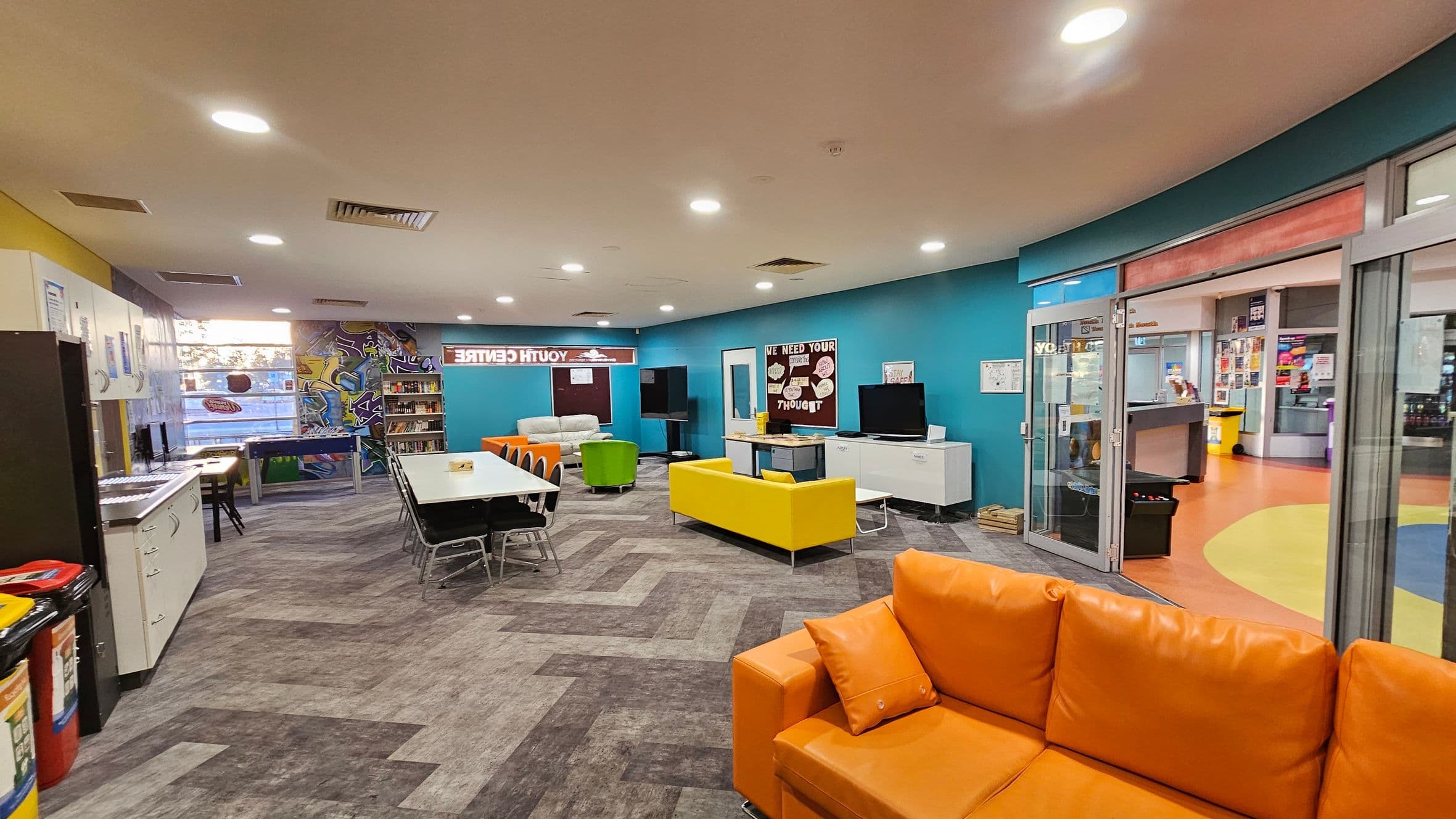 A spacious room with a vibrant, modern design. On the left, there are white cabinets and a row of computers. In the centre, a long table with black chairs is surrounded by colourful seating, including a yellow sofa and an orange couch. The back wall features a graffiti-style mural and bookshelves. On the right, glass doors lead to another area with an orange and yellow floor. The ceiling is fitted with recessed lighting, and the floor has a grey, patterned design.
