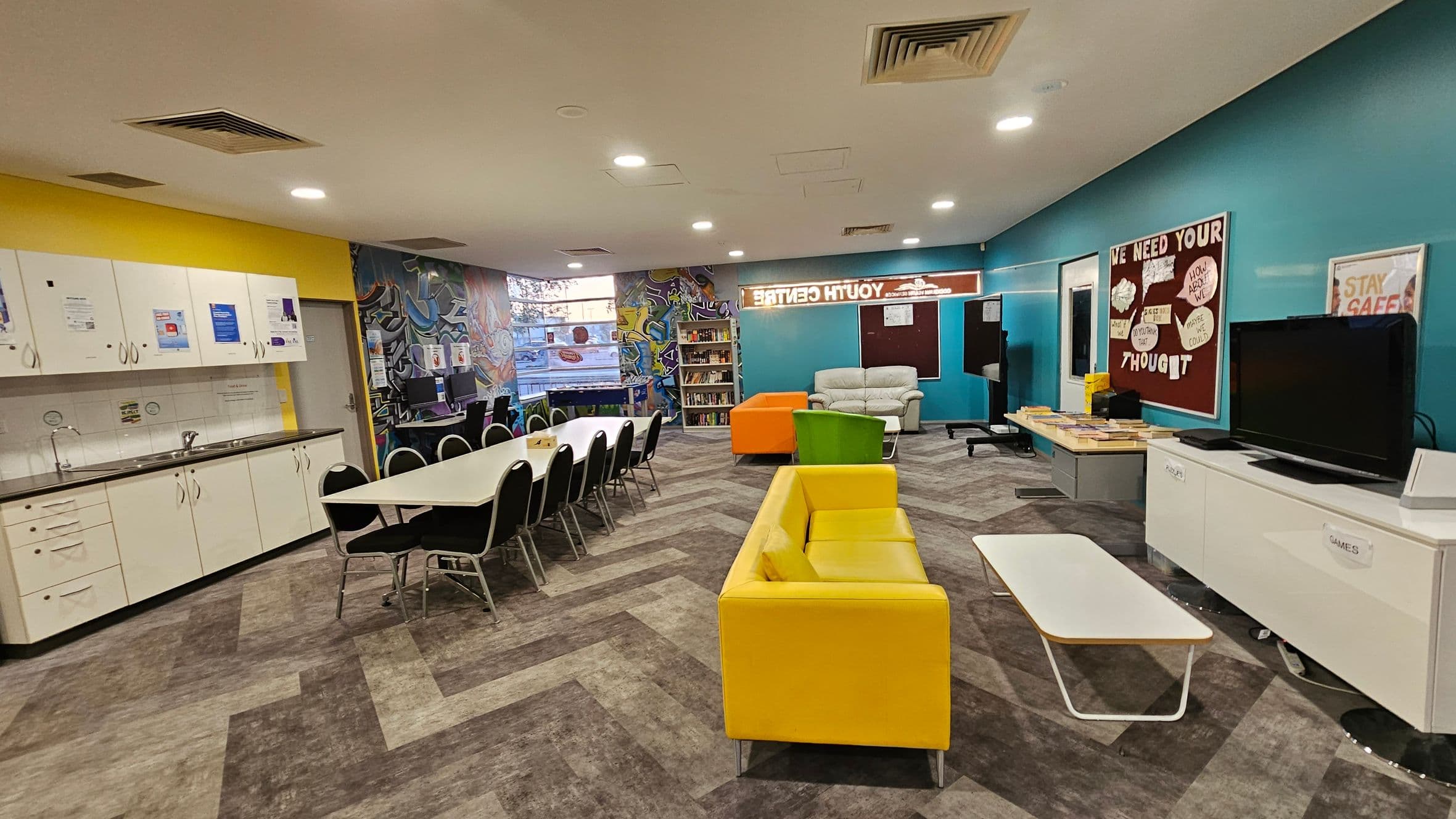 A brightly lit room with a yellow and blue colour scheme. On the left, there is a kitchenette with white cabinets and a sink. In the centre, a long table with black chairs is set up. The right side features a yellow sofa, a green chair, and a white coffee table. A TV is placed on a cabinet labelled "GAMES". The back wall has a bulletin board with various messages and a bookshelf. The floor has a patterned design, and the ceiling has recessed lighting.