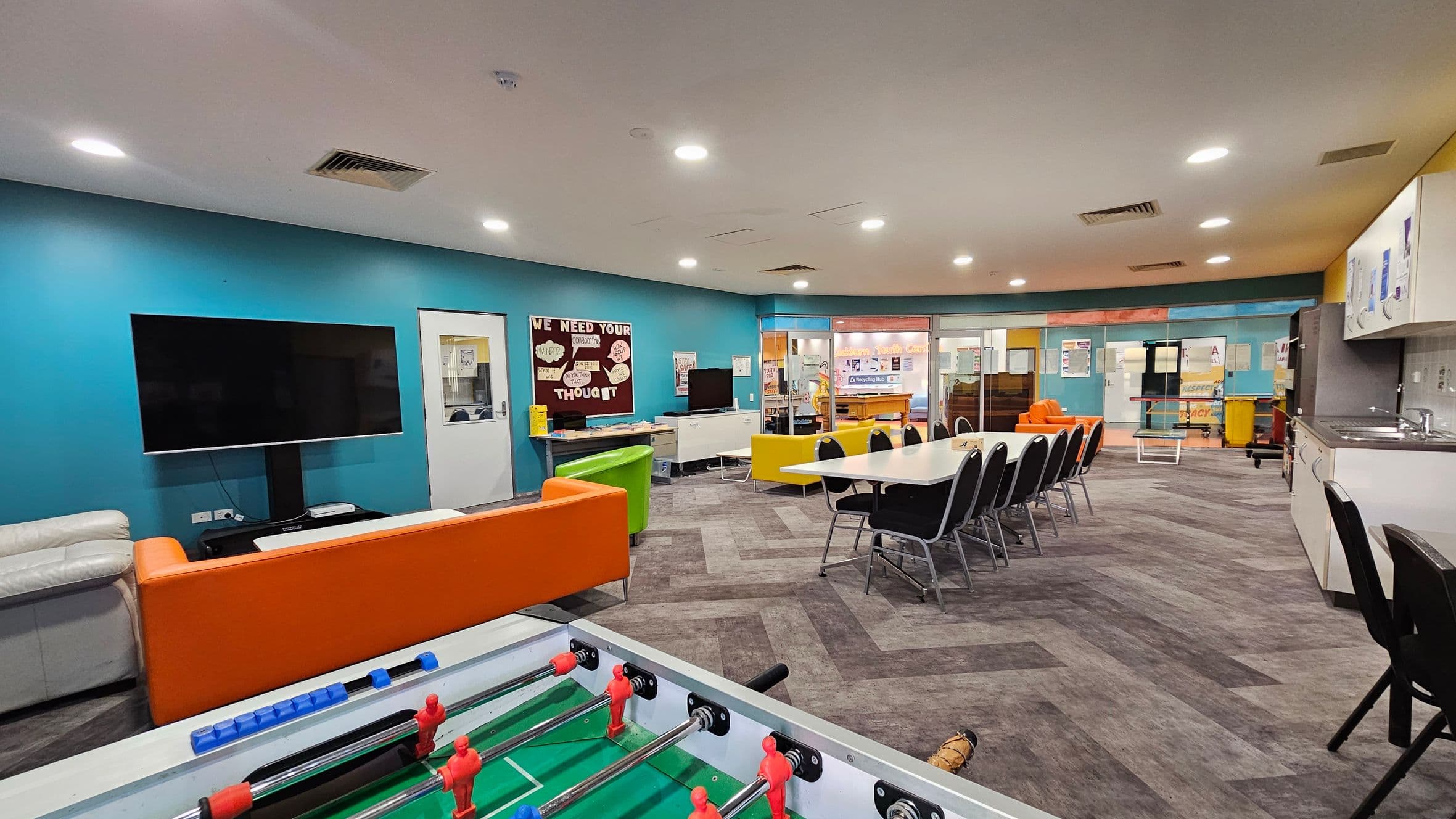 A spacious room with a foosball table in the foreground. On the left, there's a large TV mounted on a blue wall, with a white and orange sofa nearby. In the centre, a long table with black chairs is set up. The background features a bulletin board and a kitchenette area on the right, with a sink and cabinets. The floor has a patterned carpet, and the ceiling is equipped with recessed lighting.