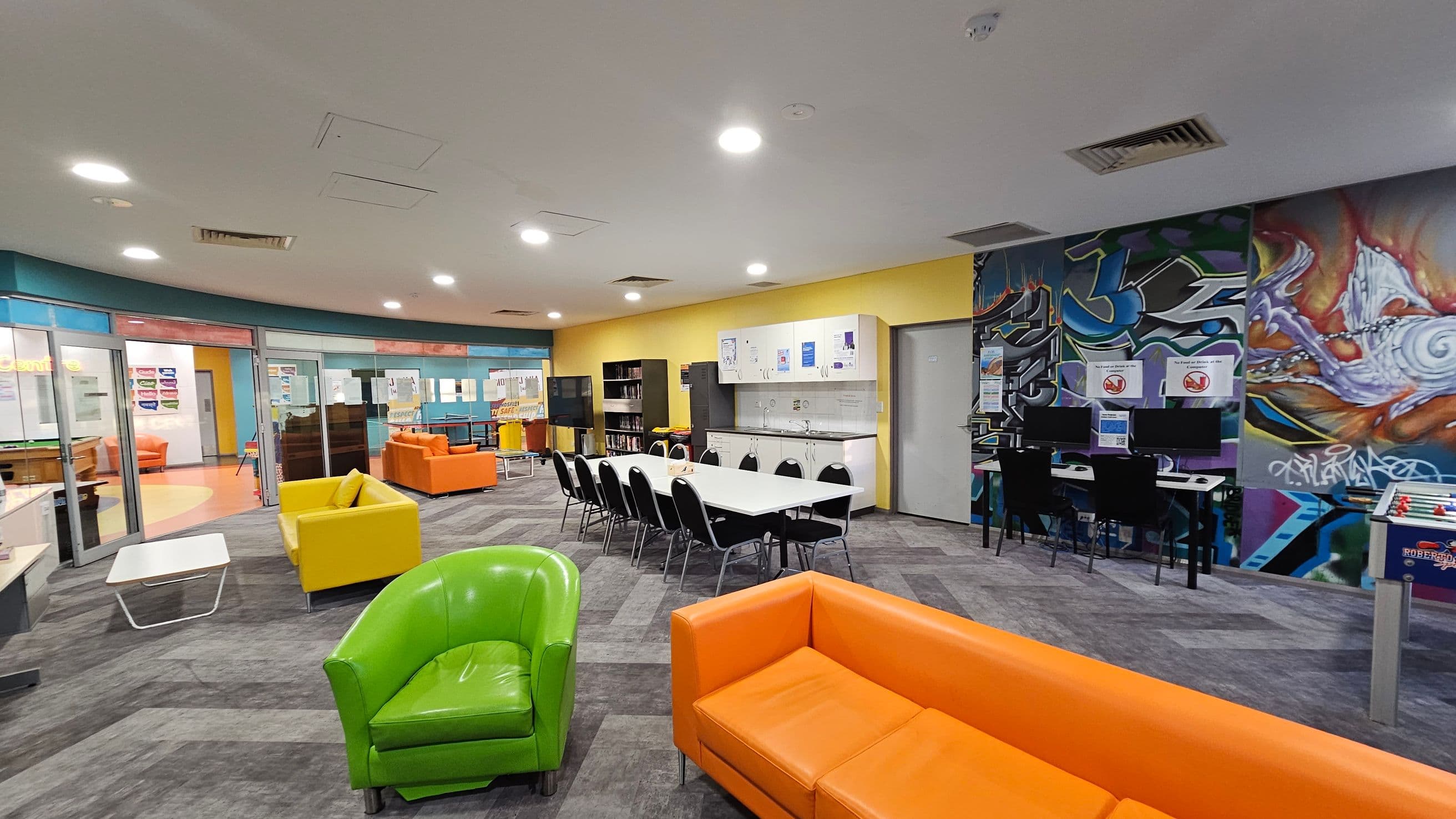 A spacious room with a modern design. On the left, there are glass doors leading to another area, with orange and yellow sofas visible. In the centre, a long white table with black chairs is set against a yellow wall with cabinets and a sink. On the right, a vibrant graffiti mural decorates the wall above computer stations. The floor is carpeted in a grey pattern, and the ceiling is white with recessed lighting. Brightly coloured sofas, including green and orange, are scattered throughout.