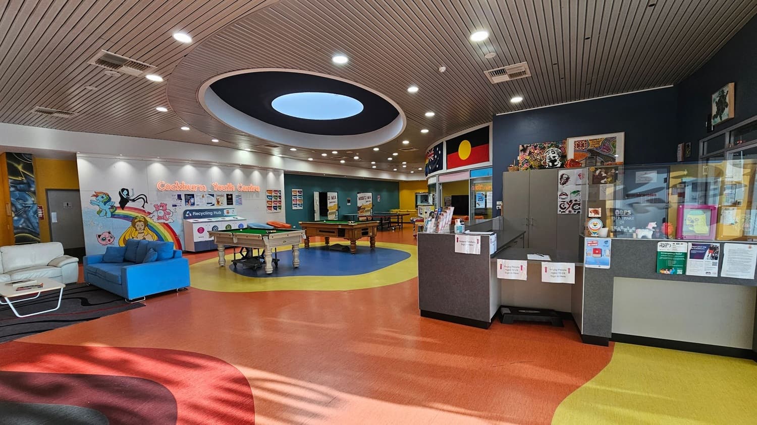 A colourful youth centre interior with a vibrant, patterned floor. On the left, there are white and blue sofas near a wall with cartoon murals and the words "Cockburn Youth Centre". In the centre, two pool tables are placed on a circular blue section of the floor. The right side features a reception area with a counter displaying brochures and posters. The ceiling is wooden with a large circular light fixture. Flags and artwork are displayed above the reception area.