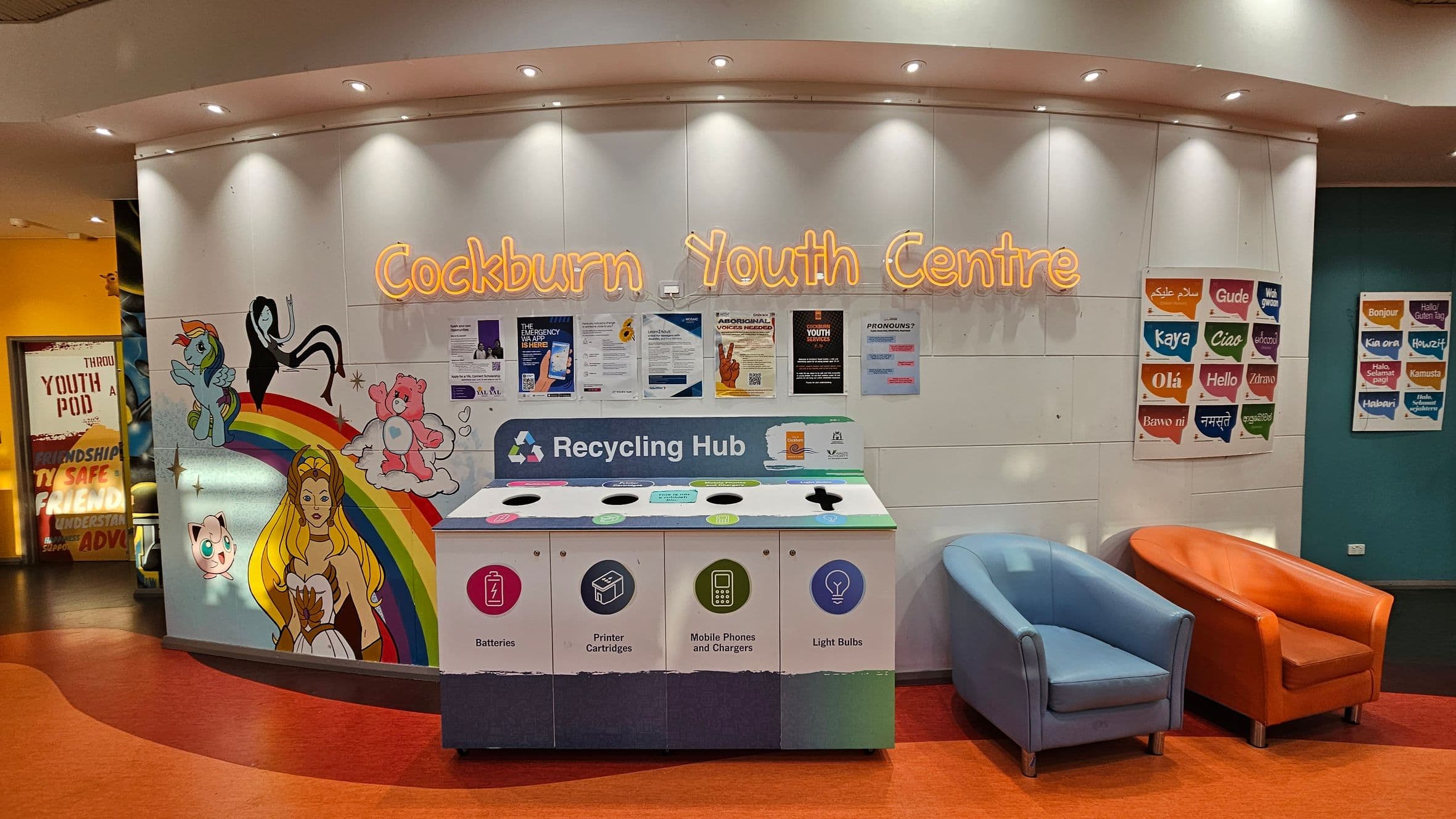 A colourful wall with the words "Cockburn Youth Centre" in neon lights. Below, a recycling hub with sections for batteries, printer cartridges, mobile phones, and light bulbs. To the left, a mural featuring cartoon characters and a rainbow. On the right, two chairs, one blue and one orange, sit on an orange floor. Posters with greetings in various languages are displayed on the right wall. The ceiling has recessed lighting.