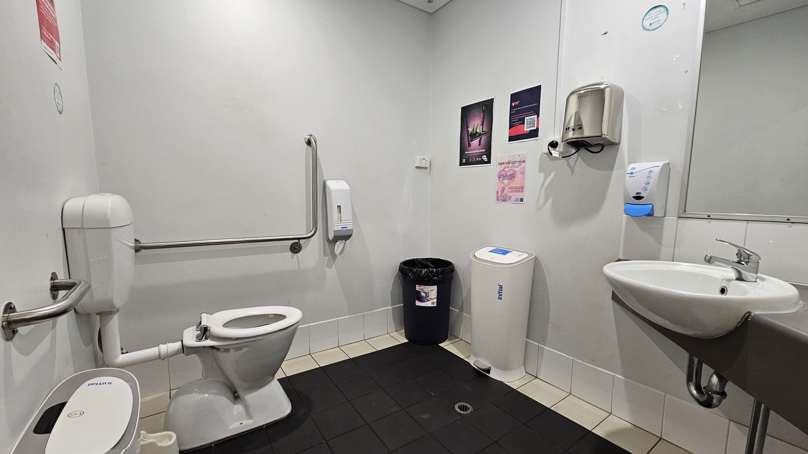 A public restroom with a white toilet on the left, equipped with a support rail. A black bin and a white sanitary bin are against the back wall. The right side features a wall-mounted sink with a silver tap, a soap dispenser, and a hand dryer above. The walls are adorned with posters. The floor is tiled in black and beige, and the ceiling is plain white with recessed lighting.