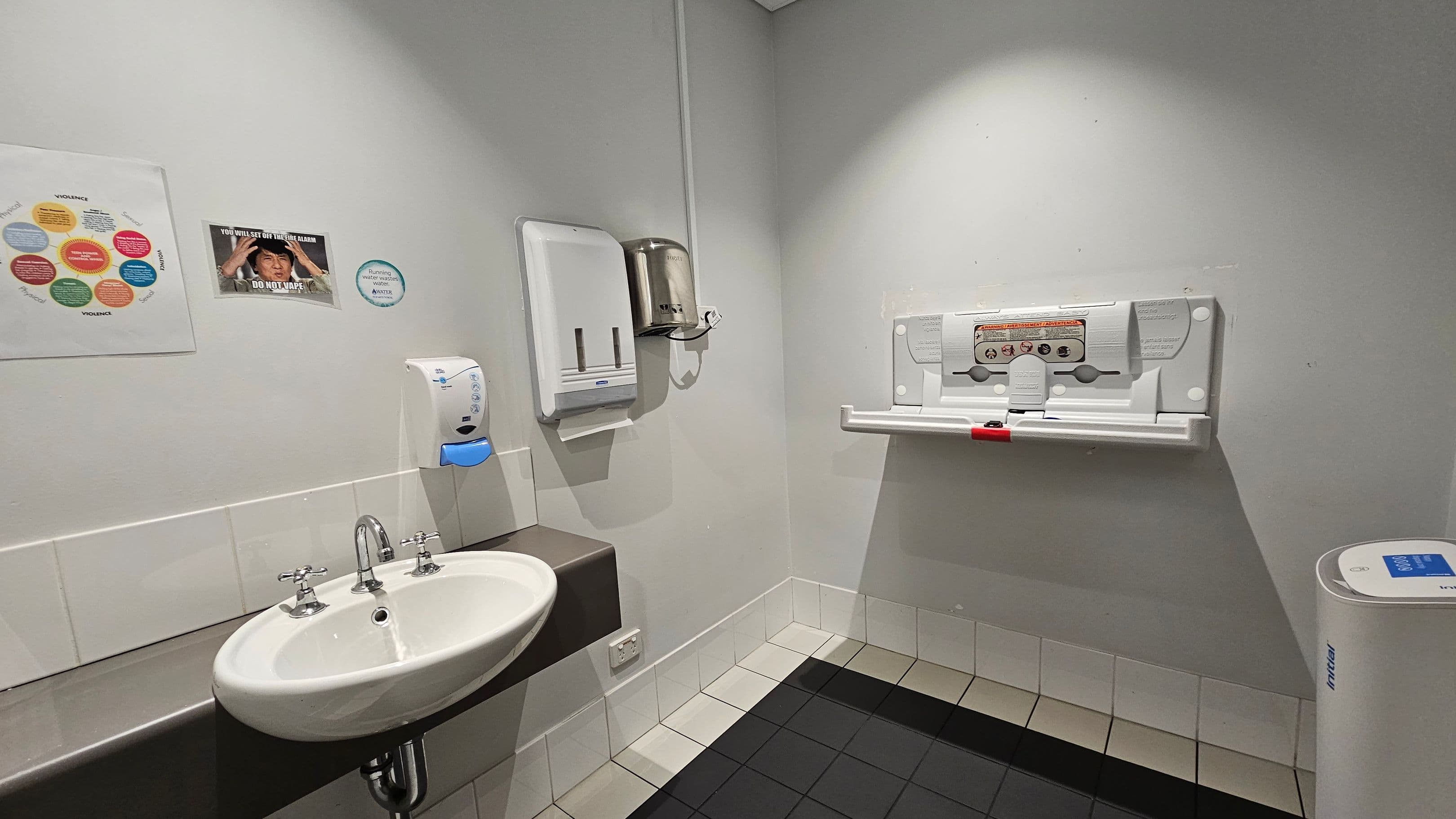 A public restroom with a white sink and silver taps on the left, mounted on a brown counter. Above the sink are a soap dispenser and a paper towel holder. On the wall, there are posters and a circular sign. To the right, a baby changing station is attached to the wall. The floor is tiled in black and light colours, and the walls are light-coloured. A white bin is in the corner on the right. The ceiling is plain and light-coloured.