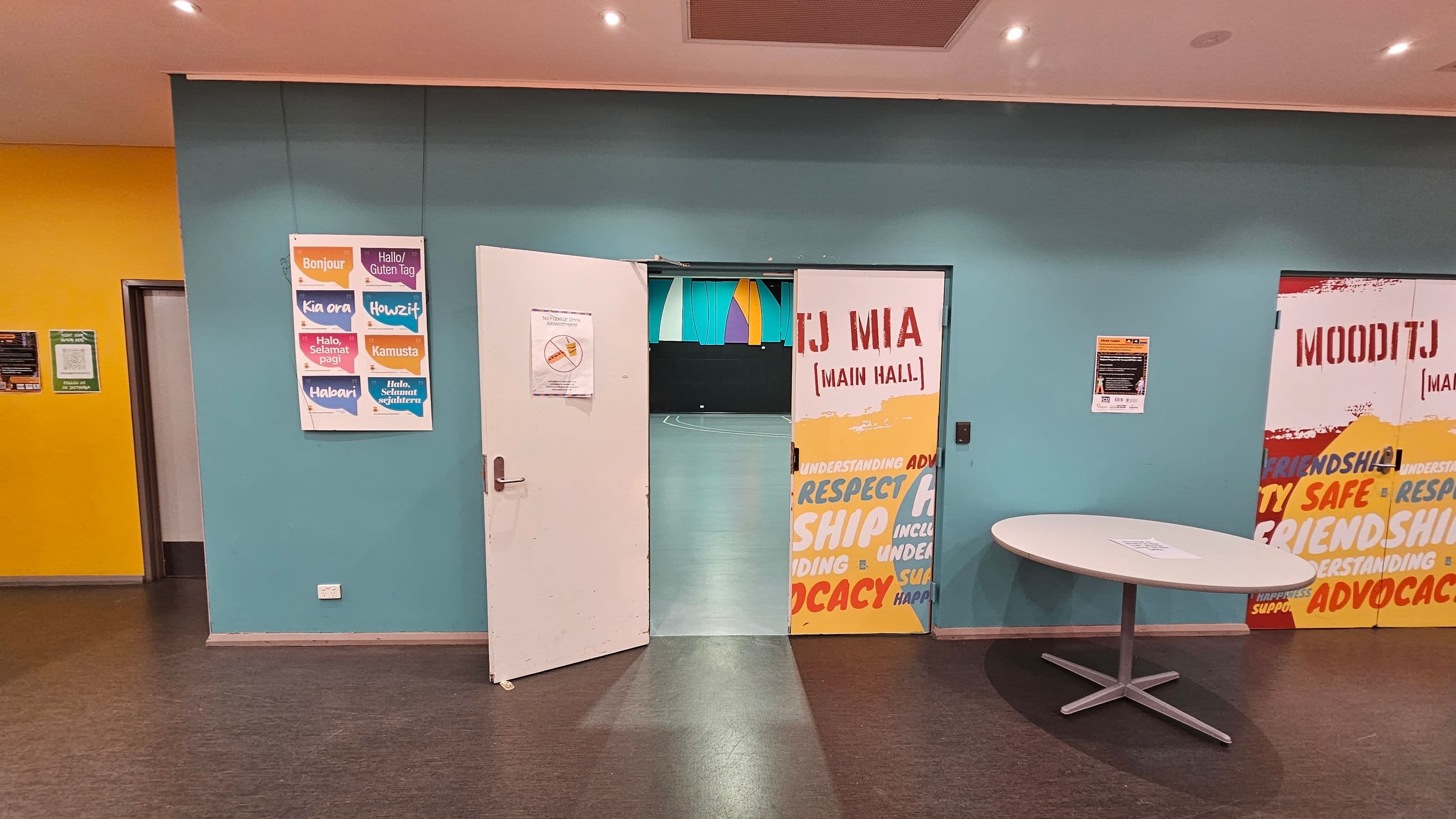 A room with a blue wall featuring a partially open door leading to a hall. The door has a sign with "TJ MIA (MAIN HALL)" and colourful words like "RESPECT" and "FRIENDSHIP." To the left, a poster displays greetings in various languages. On the right, a round table with a paper on it. The floor is dark, and the ceiling has recessed lighting. The left wall is yellow with a small doorway.