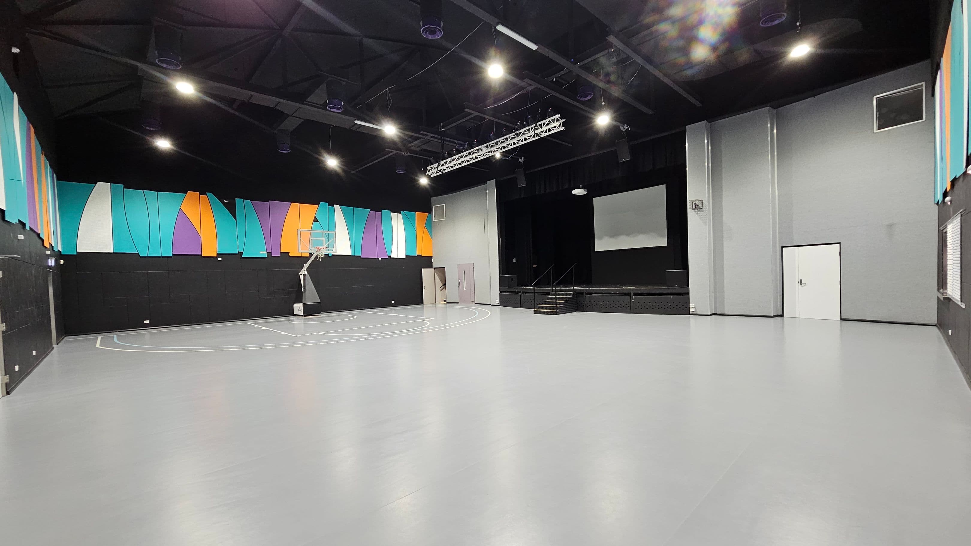 A spacious indoor gymnasium with a smooth grey floor and a high, dark ceiling. On the left, colourful geometric panels in blue, orange, and purple adorn the upper walls. A basketball hoop is positioned near the left wall. In the centre, a stage with stairs is visible, featuring a large screen above it. On the right, a plain grey wall with a white door and a small window. Bright overhead lights illuminate the space.