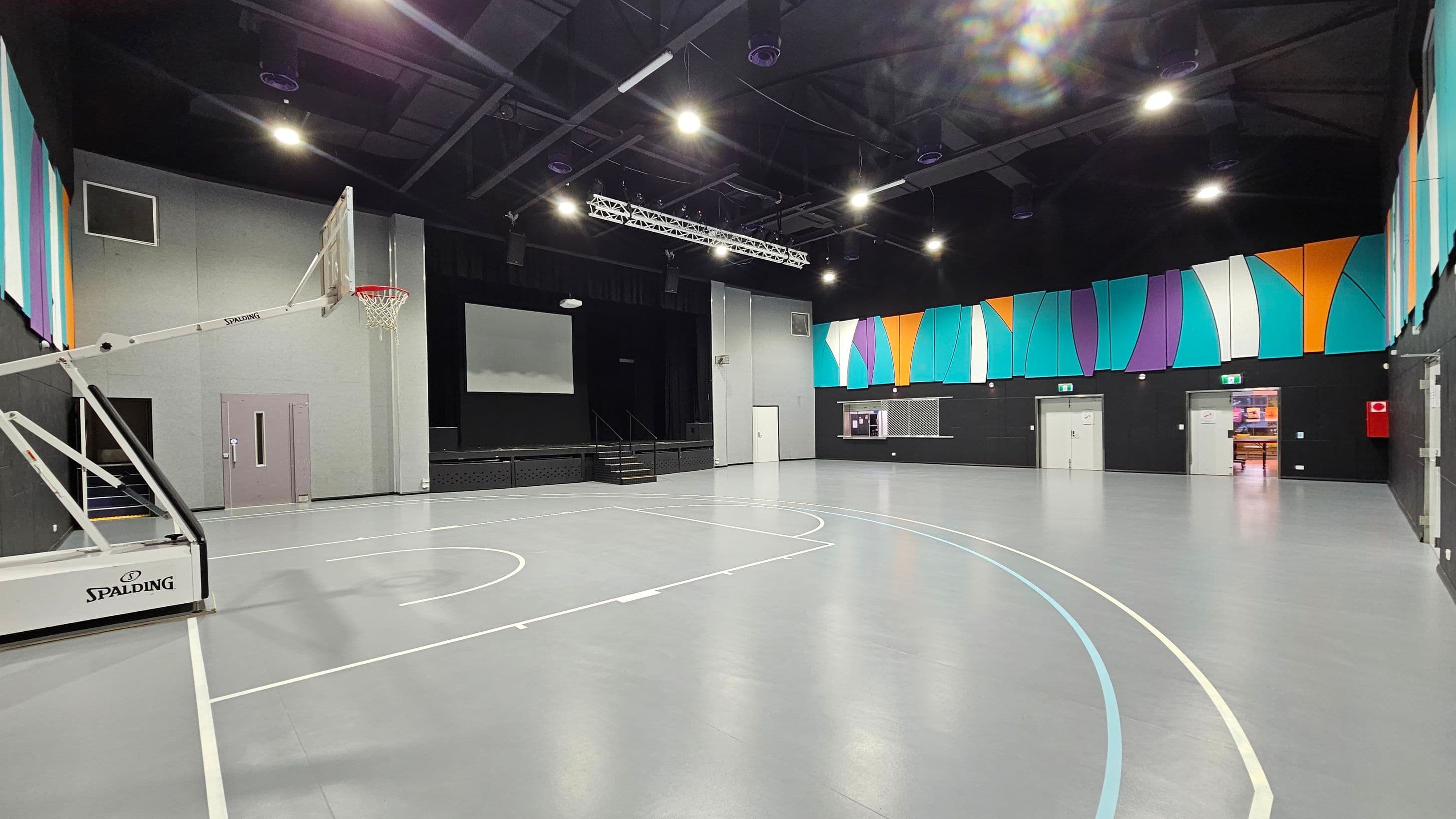 A spacious indoor gymnasium with a grey floor marked for basketball. On the left, a basketball hoop with a "Spalding" logo is visible. The back wall features a stage with a large screen and steps leading up to it. The right wall is adorned with colourful geometric panels in teal, orange, and purple. Several doors are present, including an open one showing a glimpse of another room. The ceiling is high with exposed beams and multiple lights.