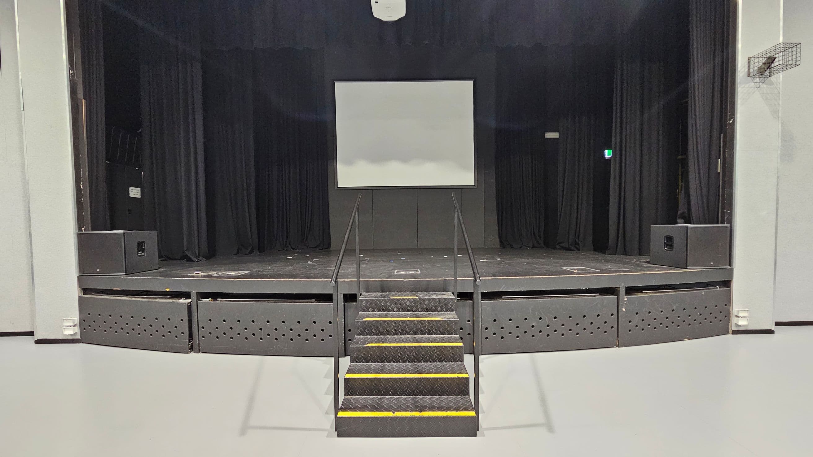 A stage with a central staircase leading up to it, featuring black railings. The stage has a large white screen in the middle, flanked by black curtains. Two large speakers are positioned on either side of the stage. The floor is light grey, and the ceiling is not visible. A small exit sign is illuminated on the right side.