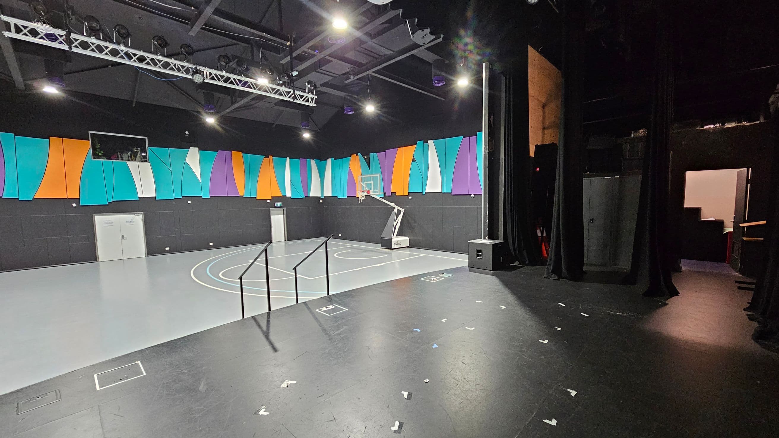 A multi-purpose indoor space with a basketball court on the left, featuring a hoop and court markings. The walls are adorned with colourful panels in orange, teal, purple, and white. The ceiling has exposed beams and lighting fixtures. On the right, there is a stage area with black curtains and a speaker. A doorway leads to a backstage area. The floor is a mix of grey and black surfaces.