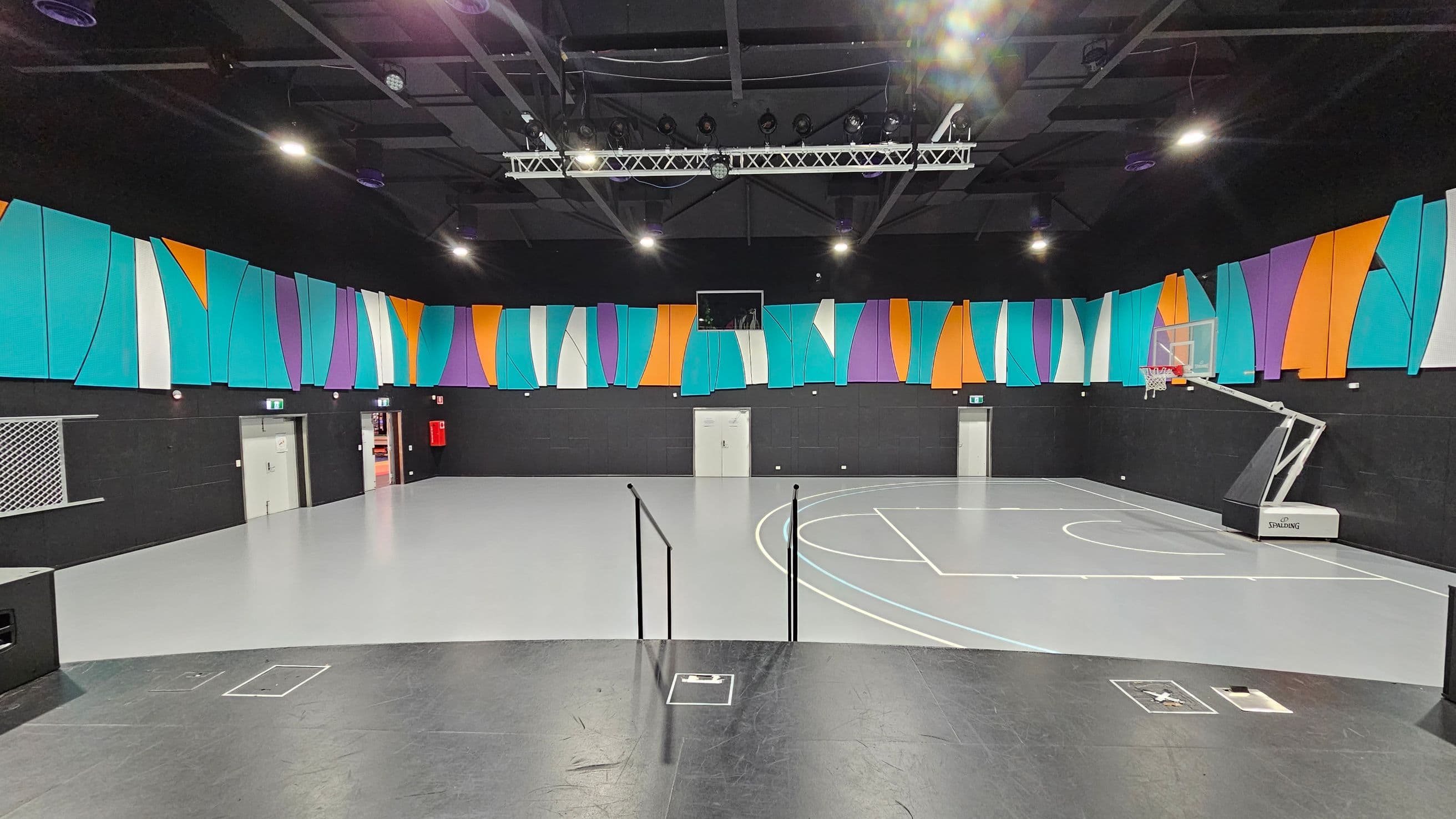 A modern indoor basketball court with a grey floor and a black ceiling. The walls are adorned with colourful, abstract panels in shades of turquoise, orange, purple, and white. A basketball hoop is positioned on the right side. There are multiple doors along the back wall, and a fire extinguisher is visible. Overhead, there are lights and a metal truss system. The foreground features a black platform with steps leading down to the court.
