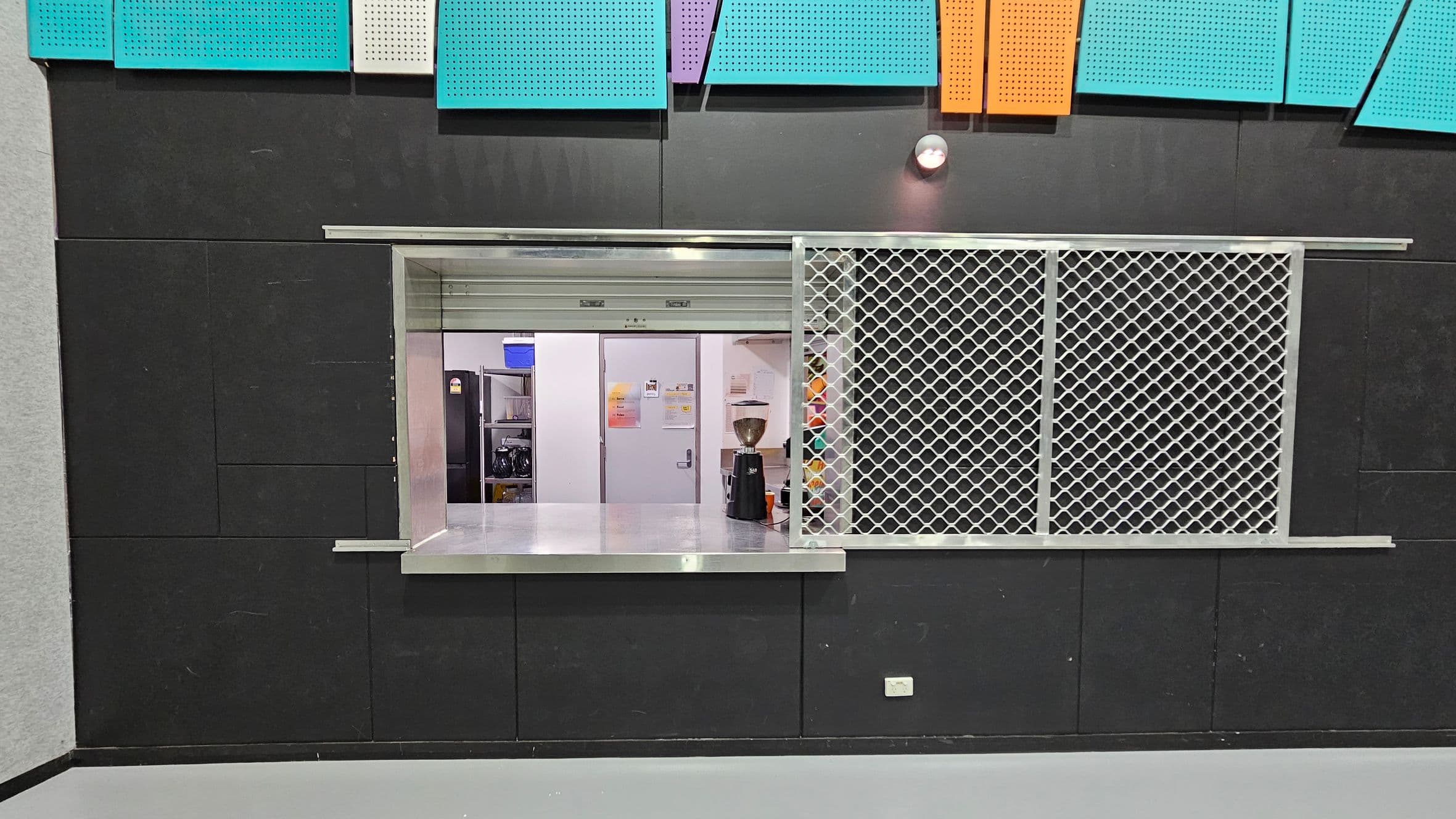 A service counter with a metal mesh screen on the right and an open window on the left. Behind the counter, there is a white door and shelves with various items, including a blue container and a coffee grinder. The wall is black with colourful acoustic panels in teal, purple, and orange at the top. The floor is grey, and there is a small light fixture above the counter.